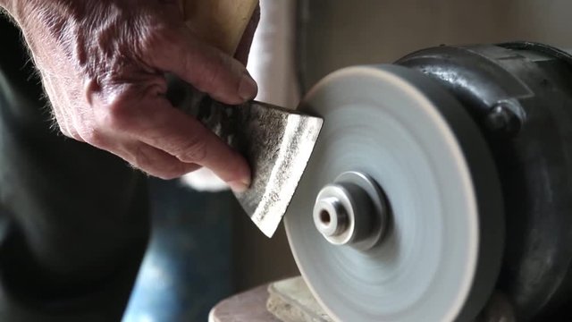 Hands hold an ax near the abrasive machine. The man sharpens the ax blade on the machine.