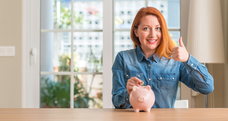 Redhead woman saves money in piggy bank at home happy with big smile doing ok sign, thumb up with fingers, excellent sign