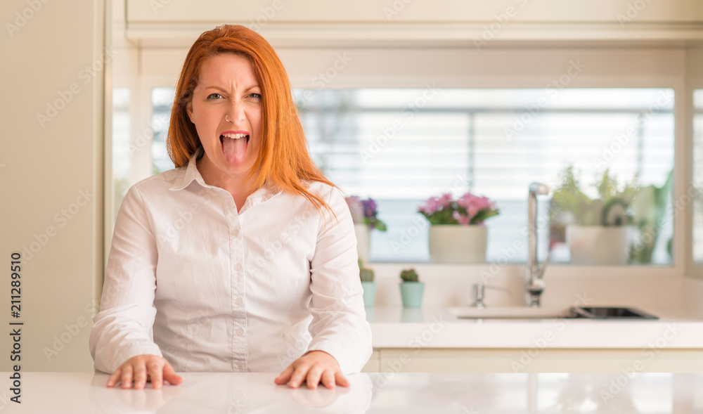 Poster Redhead woman at kitchen sticking tongue out happy with funny expression. Emotion concept.