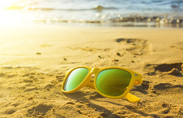 Fototapeta na wymiar Yellow style sunglasses on sandy beach in summer. Sun and sea waves in the background, depth of field focus blur