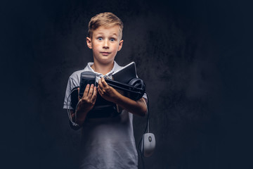 Cute schoolboy dressed in a white t-shirt, holds full digital set for entertainment at a studio. Isolated on dark textured background.