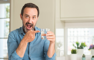 Middle age man drinking a glass of water very happy pointing with hand and finger