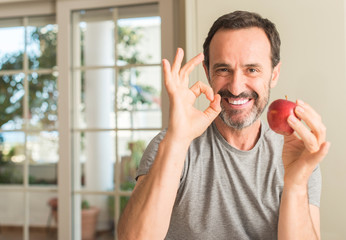 Middle age man eating healthy red apple doing ok sign with fingers, excellent symbol