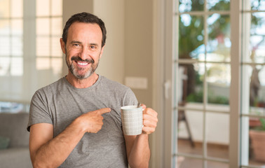 Middle age man drinking coffee in a cup with surprise face pointing finger to himself