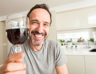 Middle age man drinking a glass of wine with a happy face standing and smiling with a confident...