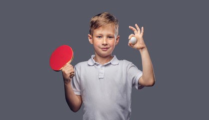 Redhead schoolboy dressed in a white t-shirt holds a ping-pong racquet and ball in a studio. Isolated on a gray background. - obrazy, fototapety, plakaty