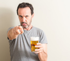 Senior man drinking beer pointing with finger to the camera and to you, hand sign, positive and...