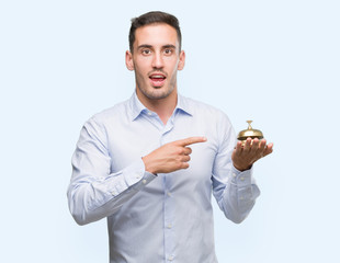 Handsome young man holding hotel ring bell very happy pointing with hand and finger