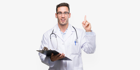 Handsome young doctor man holding a clipboard surprised with an idea or question pointing finger...