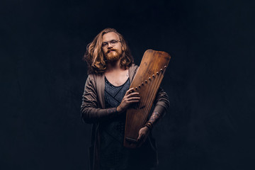 Redhead hipster male with long luxuriant hair and full beard dressed in casual clothes holds a Russian traditional musical instrument - gusli. Isolated on the dark background.