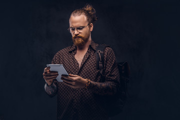 Portrait of a redhead hipster student in glasses dressed in a brown shirt, holds a backpack and digital tablet, posing at a studio. Isolated on a dark background.
