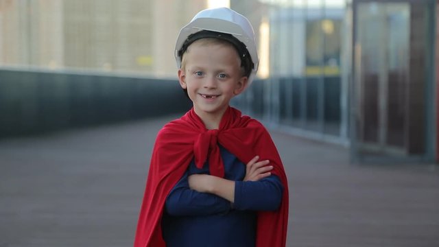 Portrai a boy in a superman costume building man, wearing a white helmet, a skyscraper background and a construction site.