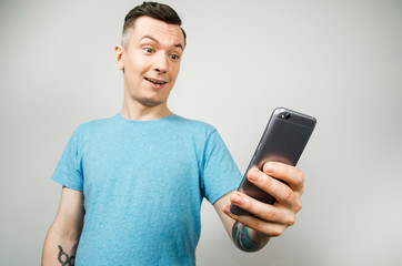 Young cheerful caucasian guy in a blue t-shirt holds a smartphone and looks at it, isolated on a light gray background.