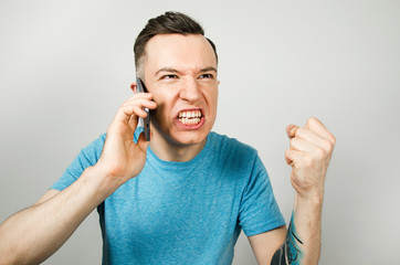 Young angry caucasian guy in blue t-shirt talking on the phone, isolated on light gray background.