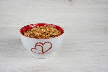 A white bowl with two hearts roasted on it full of granola on white wood table