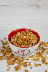 A white bowl with two hearts roasted on it full of granola on white wood table