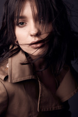 Dark close up portrait of beautiful sensual woman with nude makeup, dark hair, wearing beige trench coat. Studio shot