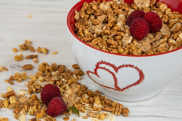 A white bowl with two hearts roasted on it full of granola whit berries on white wood table,close-up