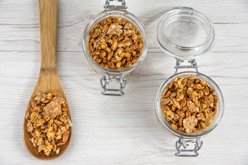 Two jars full of granola  and a wood spoon full whit granola on the white wood table, top view