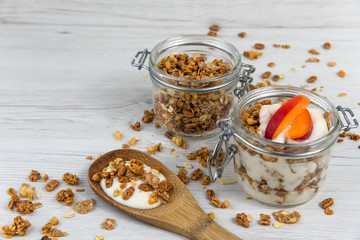 A  jar filled with granola and yogurt, wooden spoon whit granola and apricot on white wood table