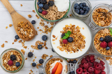 Jars full with granola, yogurt and fresh berries and wood spoon full whit granola on white wood table, top view, selective focus