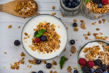 Jars full with granola, yogurt and fresh berries and wood spoon full whit granola on white wood table, top view, selective focus