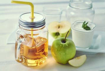 Mason Mug with apple juice and a yellow straw. One glass jar of homemade yogurt with mint and sliced apples on a white plate near window in the morning.