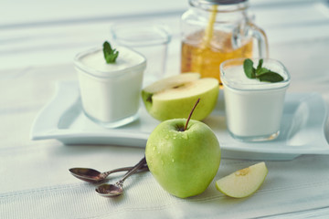 Green fresh apple with two glass jars of homemade yogurt with mint and sliced apples on a white plate near window in the morning. Mason Mug with apple juice and a straw.