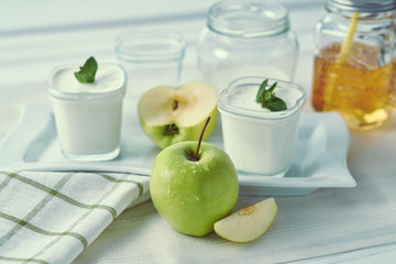 Two glass jars of homemade yogurt with mint and sliced apples on a white plate near window in the morning. Mason Mug with apple juice and a straw.