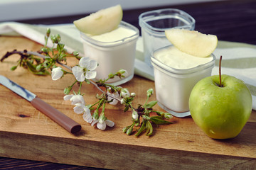 Two jars with yogurt, decorated with apple slices, on a wooden board with a knife and a branch of a blossoming apple tree.