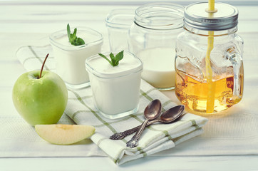 Two glass jars of homemade yogurt with mint and sliced apple on a light textile background near window in the morning. Mason Mug with yellow apple juice and a straw.