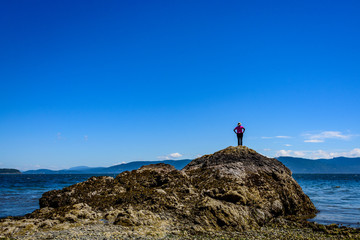 Woman in black pants and pink shirt climbing a rock formation on a beach in the San Juan Islands, the Salish Sea and other islands in the background, sunny day with a blue sky and while clouds
