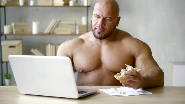 bald male bodybuilder writes text on a laptop and does not violate the diet eats a triple Burger with beef Patty