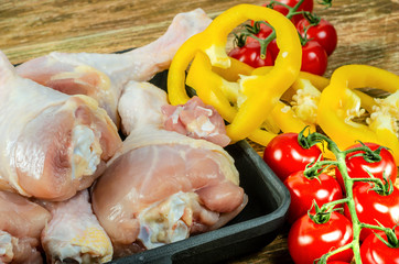 Raw organic chicken drumsticks with fresh vegetables lies in the black frying pan, on a wooden background before preparation. Cooking concept.