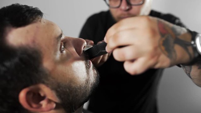 Tattoed barber shears the customer's mustage by using trimmer and comb at the barber shop, man's haircut and shaving at the hairdresser, shaving salon