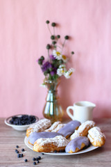 Berry glazed eclairs with blueberries on a wooden background with a decor consisting of a vase with wildflowers and a jug for milk and plate with blueberries