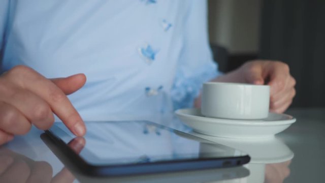 A young girl sitting at a table with a Cup of coffee, a digital tablet in her hands, leafing through the catalog of the online store on the touch screen of the tablet, cropped image.