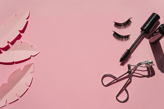 Black false lashes strips,mascara, curler on pink background with paper leaves