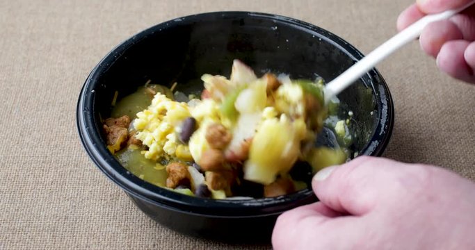 Video of a partially microwaved scrambled egg burrito meal in a black tray being stirred with a fork.