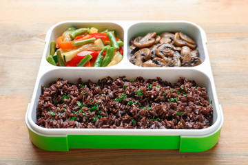 Lunch box with red rice, vegetables and mushrooms on table