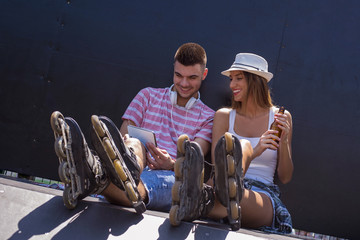 Two young people in roller skates are sitting in a skate park and using tablet