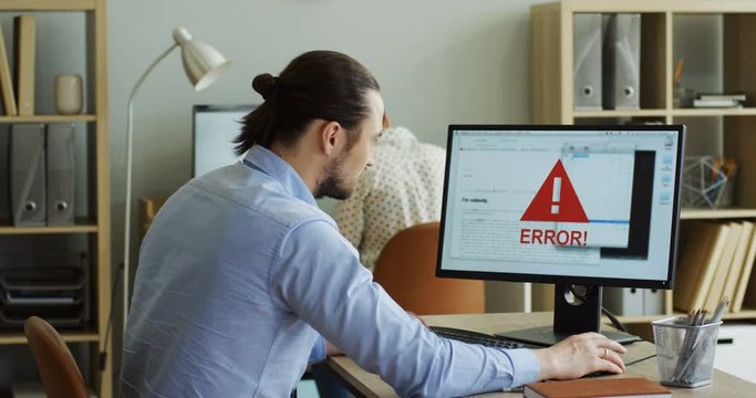 Caucasian young man sitting at his office desk and using laptop computer wich being broken and there being error on the screen, then taking his smartphone and using it. Inside.