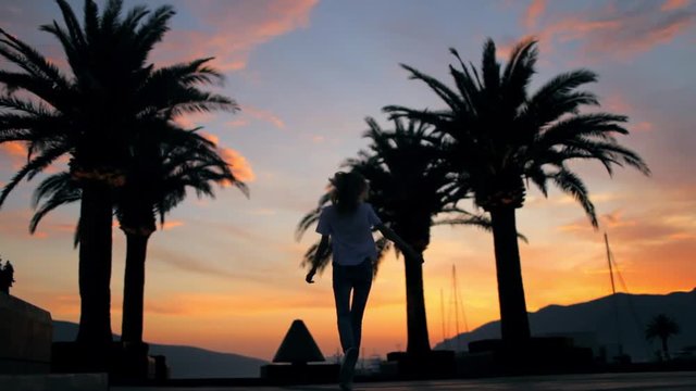silhouette of a cheerful girl walking on the waterfront with palm trees at sunset