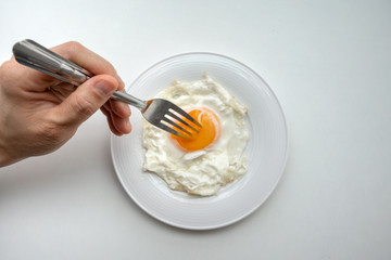 Hand holding fork to prick the egg yolk of beautiful fried egg in white plate isolated on white