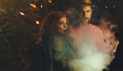 Loving couple on date. Guy and girlfriend smoke electronic cigarettes. Clouds of smoke. Red-haired girl. Man in hat and t-shirt. Lights of night city shine.