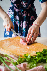 Woman hands cuts raw red meat into pieces with sharp knife on wooden board.