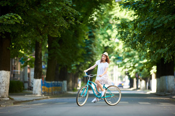 Positive pretty girl in white dress and straw hat is happy riding blue bike down wide beautiful park alley with trees around on sunny summer day. Beautiful female ride enjoying happiness healthy