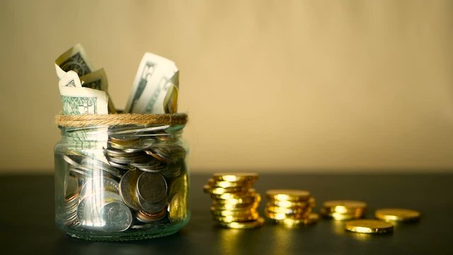 Saving money coin in jar. Symbol of investing, keeping money concept. Collecting cash banknotes in glass tin as moneybox. American dollars close-up with increasing columns of gold coins on black table
