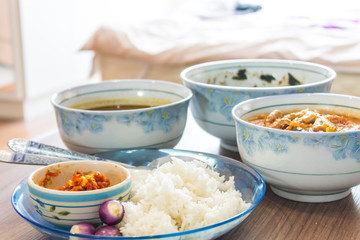 Side view inside the bedroom Pure white jasmine rice in blue plate Have a cup of red chili paste. And there are three folk red curry paste on a brown wooden table.