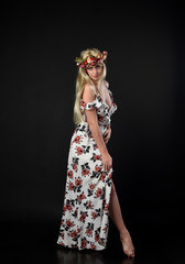 full length portrait of blonde girl wearing floral dress and a flower crown. standing pose against a black studio background.